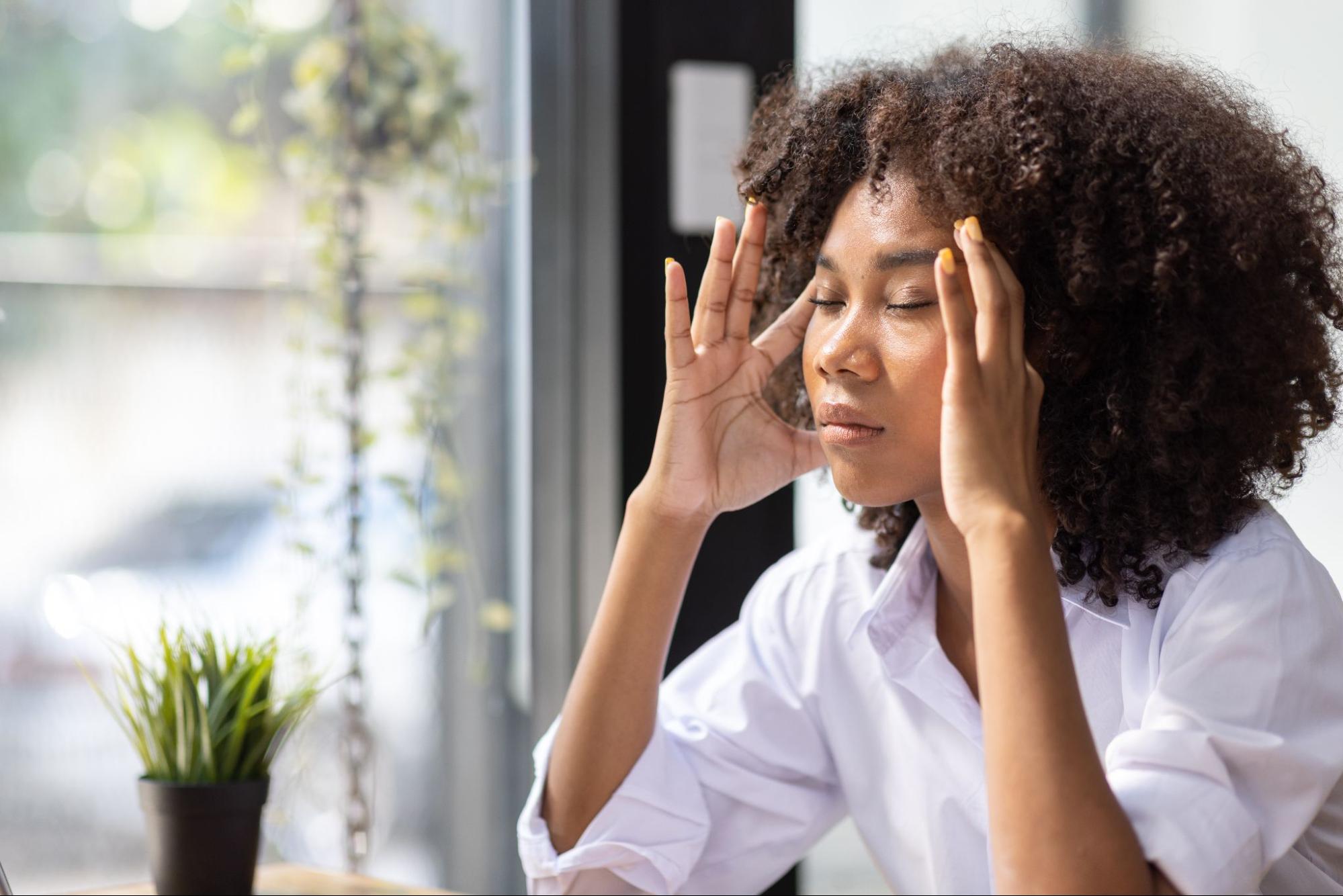 Woman at a desk with eyes closed and hands at her temples, showing stress or a headache during the menopause transition.