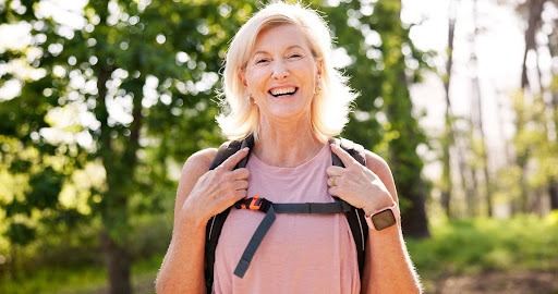 Smiling mature woman wearing a backpack in the forest, promoting bone health through physical activity like weighted vest walking for menopause.