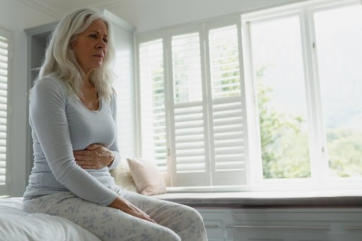 Older woman sitting on a bed holding her abdomen, representing pelvic or vaginal discomfort during menopause.