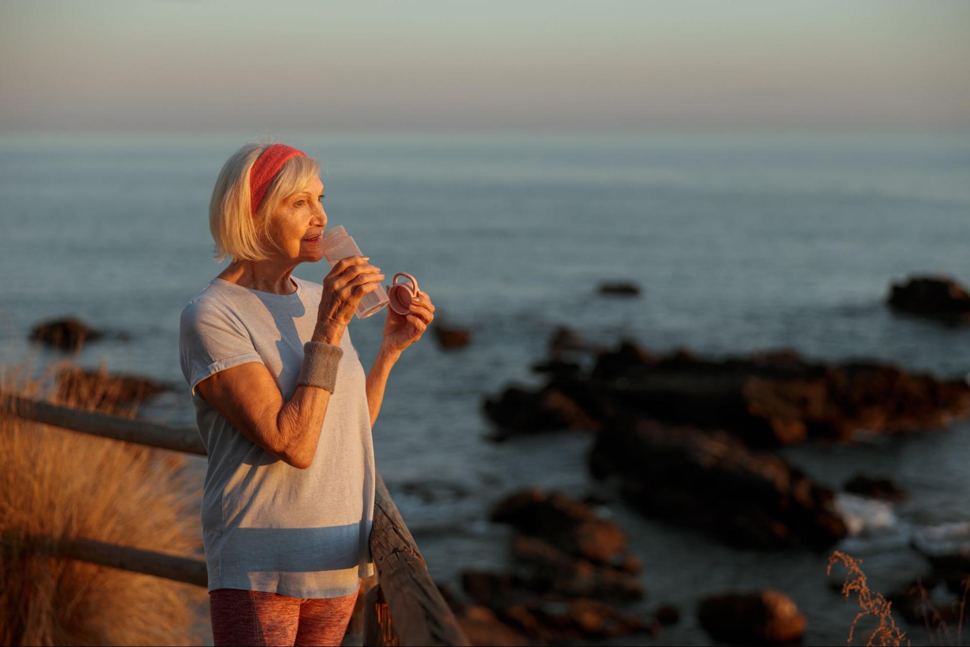 Mature woman drinking water outdoors by the ocean, supporting hydration and healthy habits during menopause.